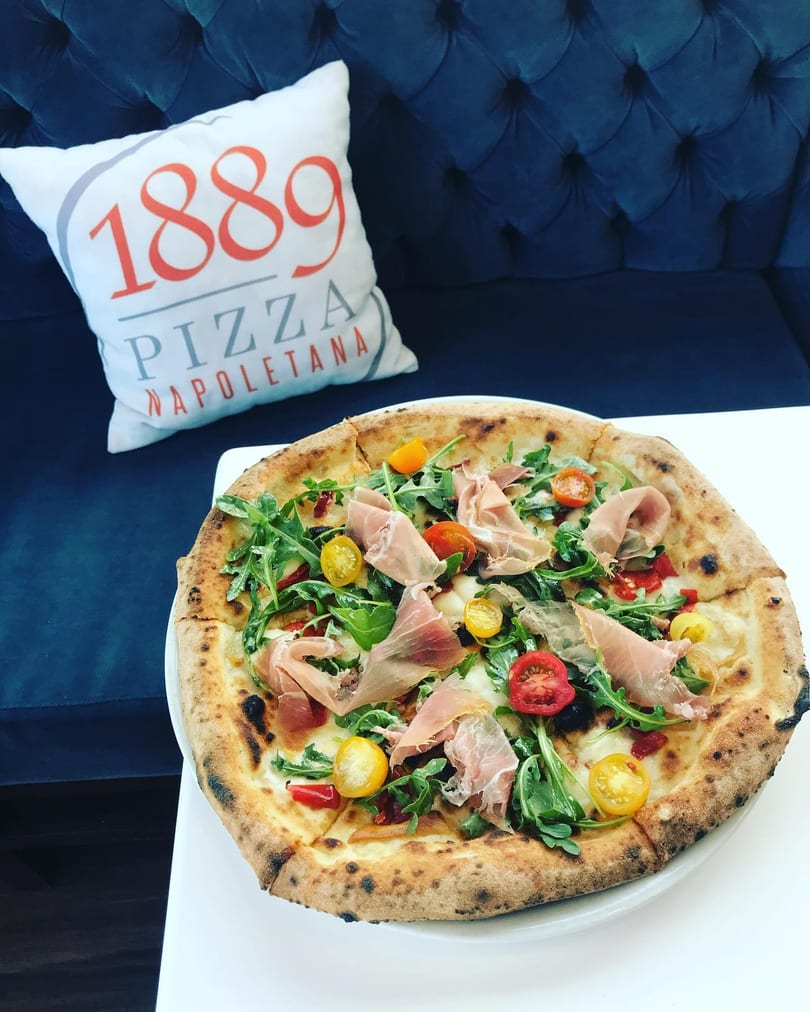A pizza with prosciutto, arugula, and cherry tomatoes on a white table next to an "1889 Pizza Napoletana" pillow.