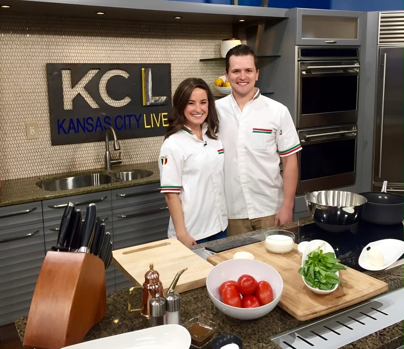Two chefs, a man and a woman, smiling in a modern kitchen with fresh ingredients on the counter.
