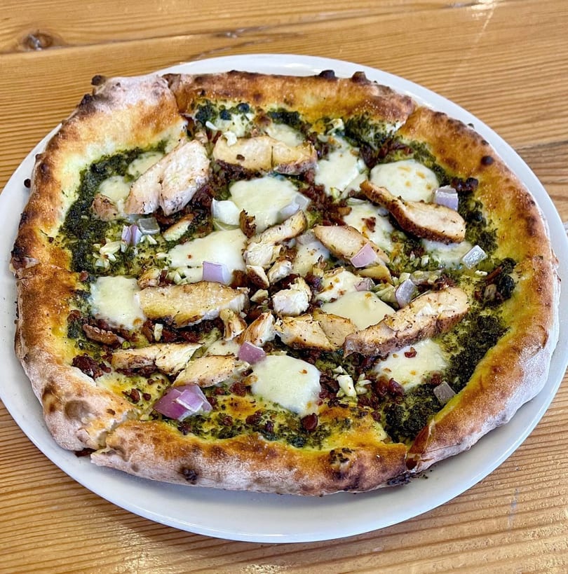 A pesto chicken pizza with mozzarella, red onion, and sun-dried tomatoes on a white plate.