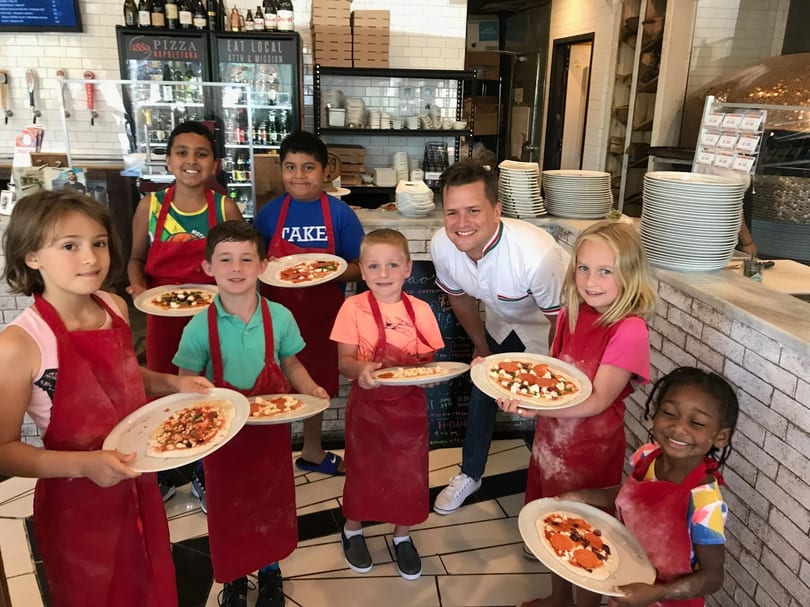 Children in red aprons proudly hold pizzas they made with a chef at 1889 Pizza Napoletana.