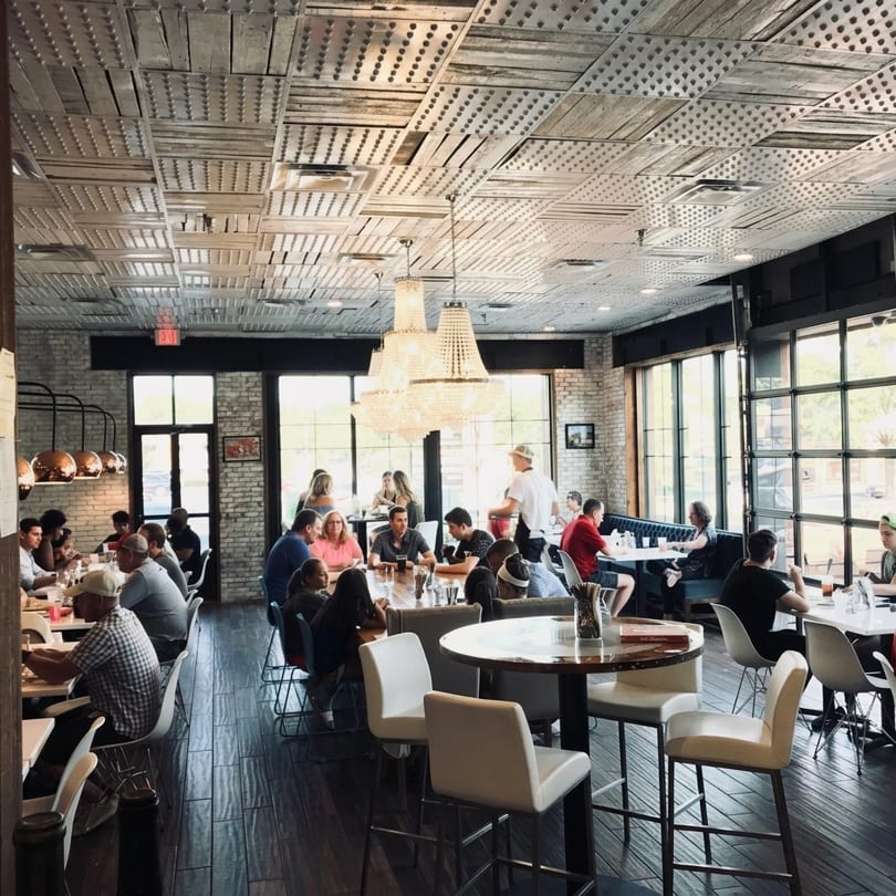 A bustling restaurant interior with patrons dining at tables under ornate chandeliers and a textured ceiling.