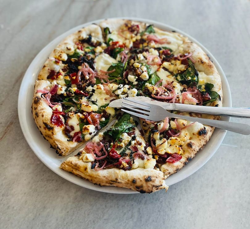 A gourmet pizza with feta, spinach, red onion, and sun-dried tomatoes on a white plate with cutlery.