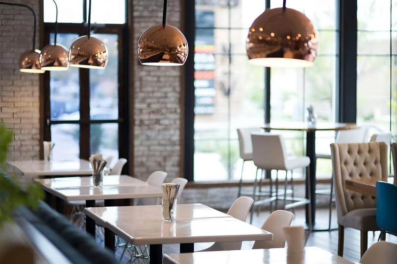 Modern restaurant interior with white tables, copper pendant lights, and large windows.