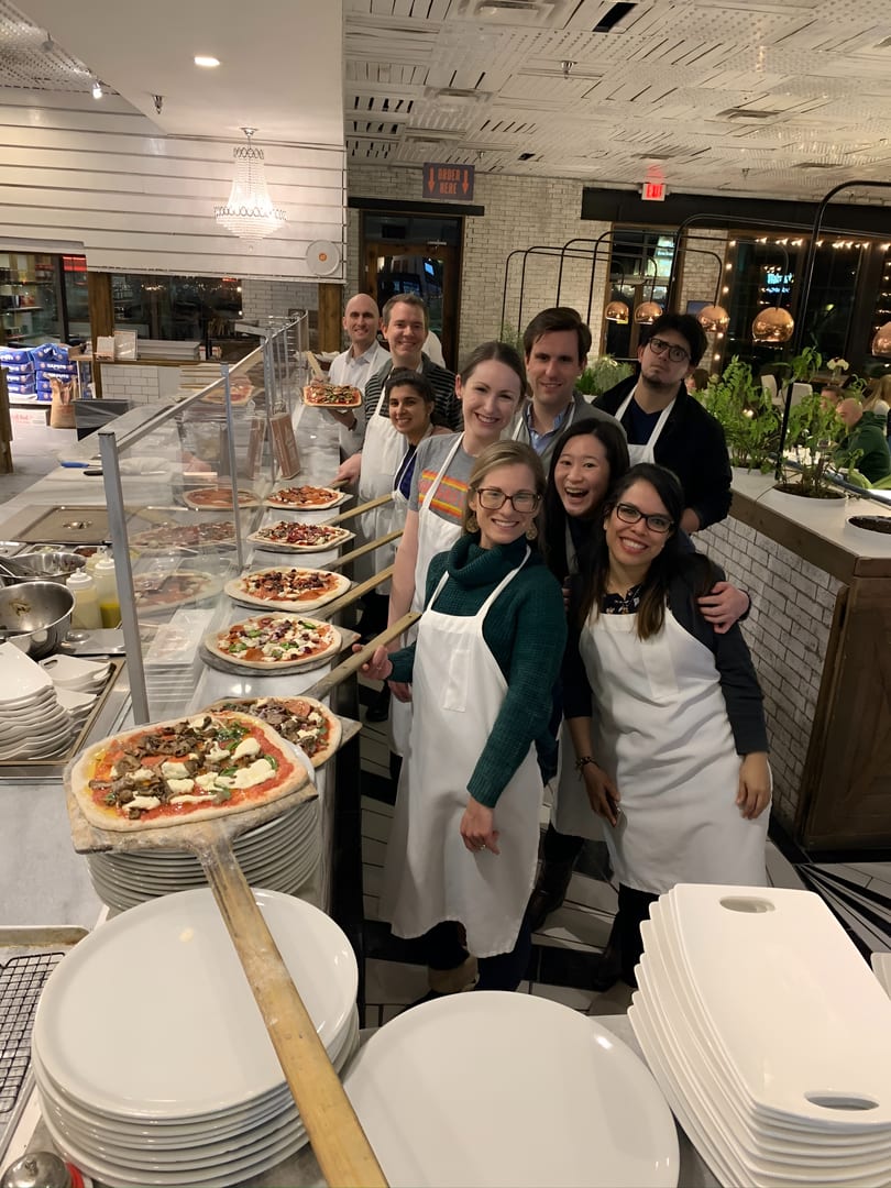 A group of smiling people in aprons hold pizza peels with uncooked pizzas in a restaurant kitchen.