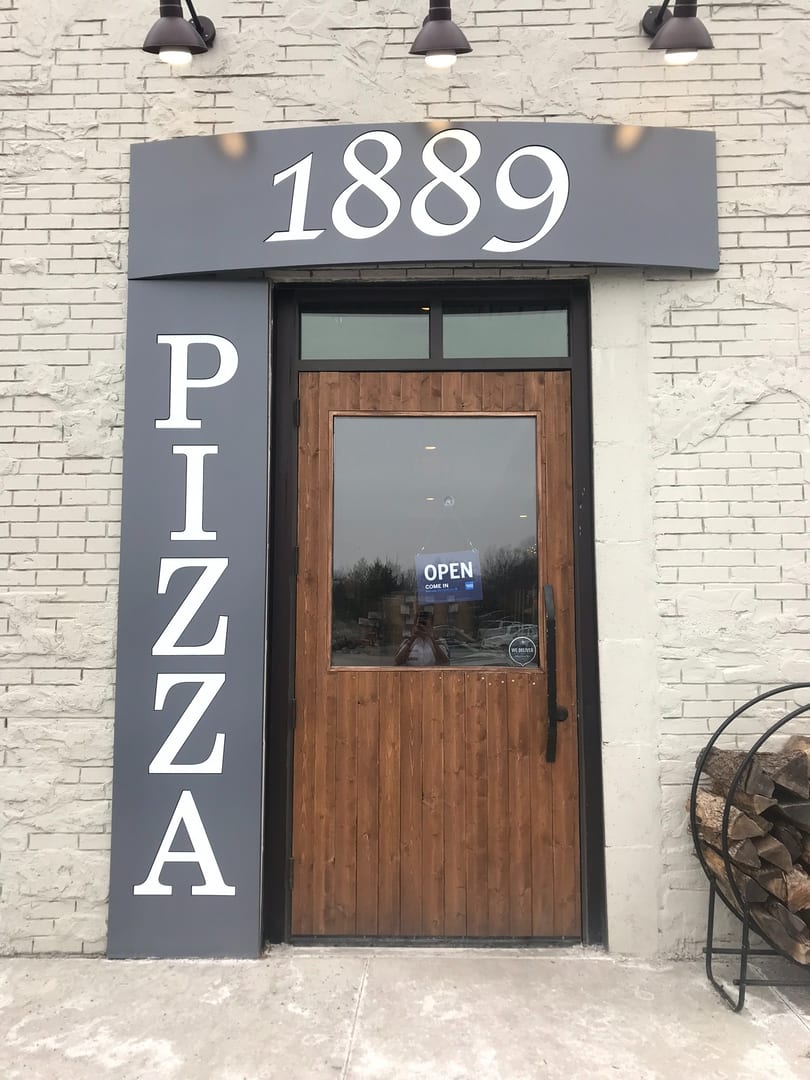 Exterior of 1889 Pizza restaurant with a wooden door, "OPEN" sign, and stacked firewood.