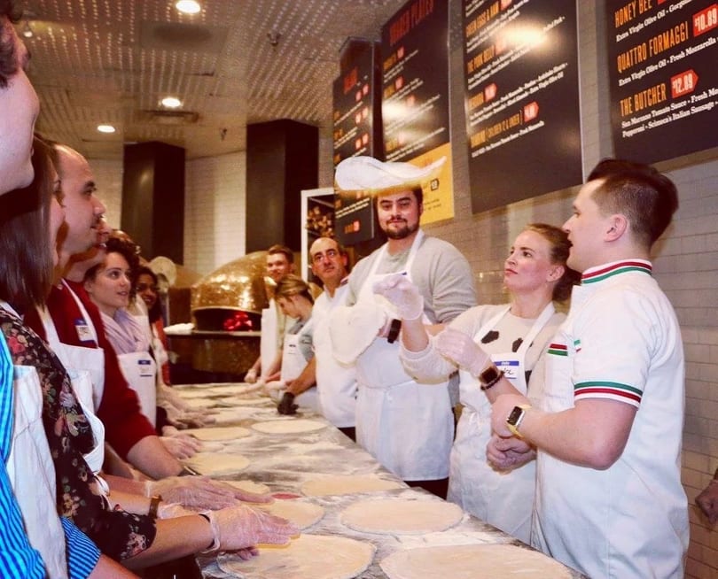 People in aprons learning to make pizza, with one person tossing dough in the air.