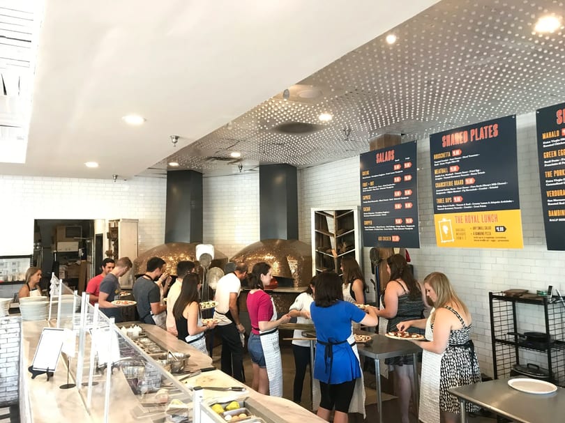 Customers line up at a counter to customize pizzas, with large ovens and menu boards in the background.