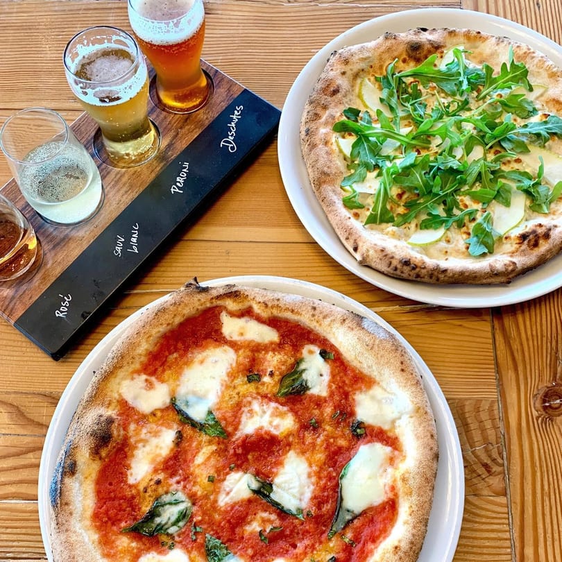 Two pizzas, one Margherita and one with arugula and pears, next to a beer and wine flight.