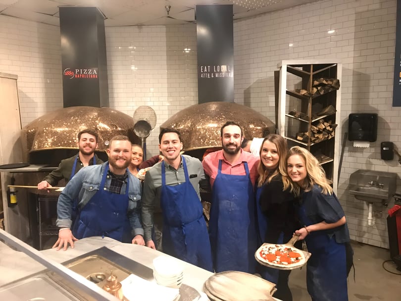 Seven smiling people in blue aprons stand in a kitchen with large pizza ovens, one holding a pizza.