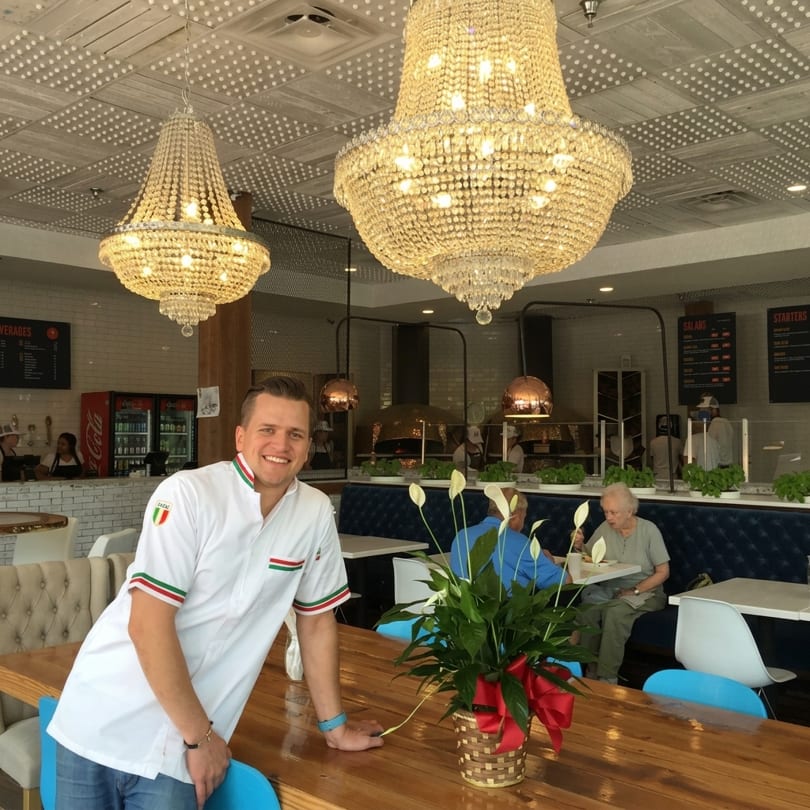 Smiling chef in a white uniform leaning on a wooden table in a bright restaurant dining room.
