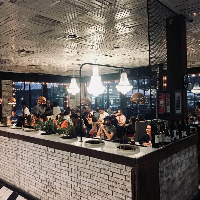 A lively restaurant interior with patrons dining at tables and a long bar with white brick.