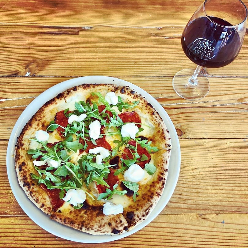Neapolitan pizza with arugula, mozzarella, and pepperoni next to a glass of red wine.